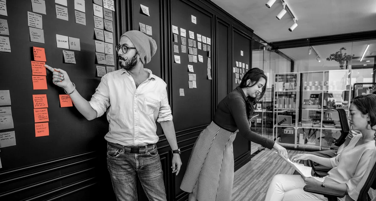 A team member pointing at one group of notes on a wall covered in written notes, while colleagues review a document